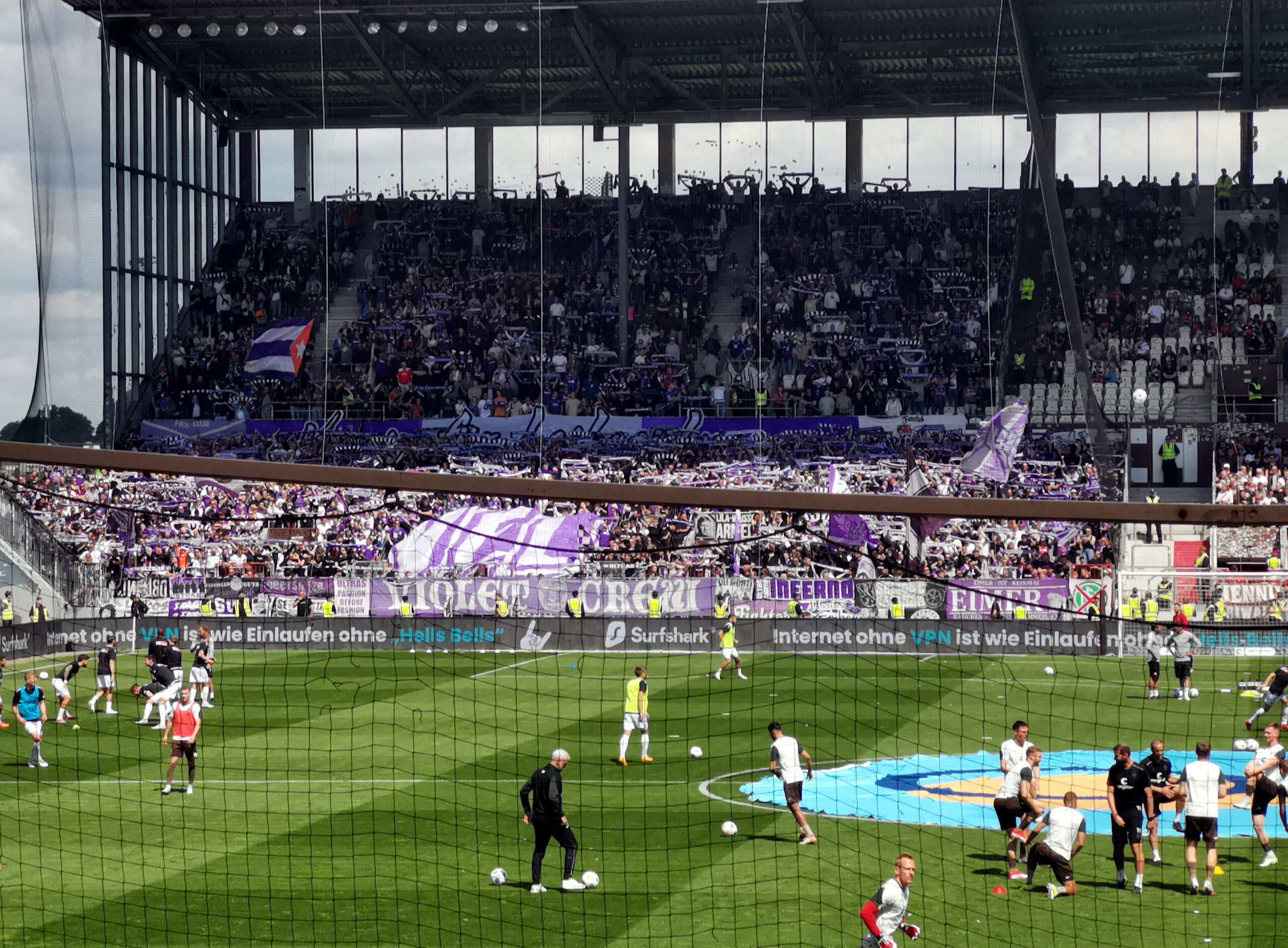 Zu sehen ist der Gästeblock im Millerntor-Stadion mit den Osnabrück-Fans, welche ein großes Banner mit ihrem Vereinswappen hochhalten. Im Vordergrund machen sich die Spieler auf dem Rasen warm